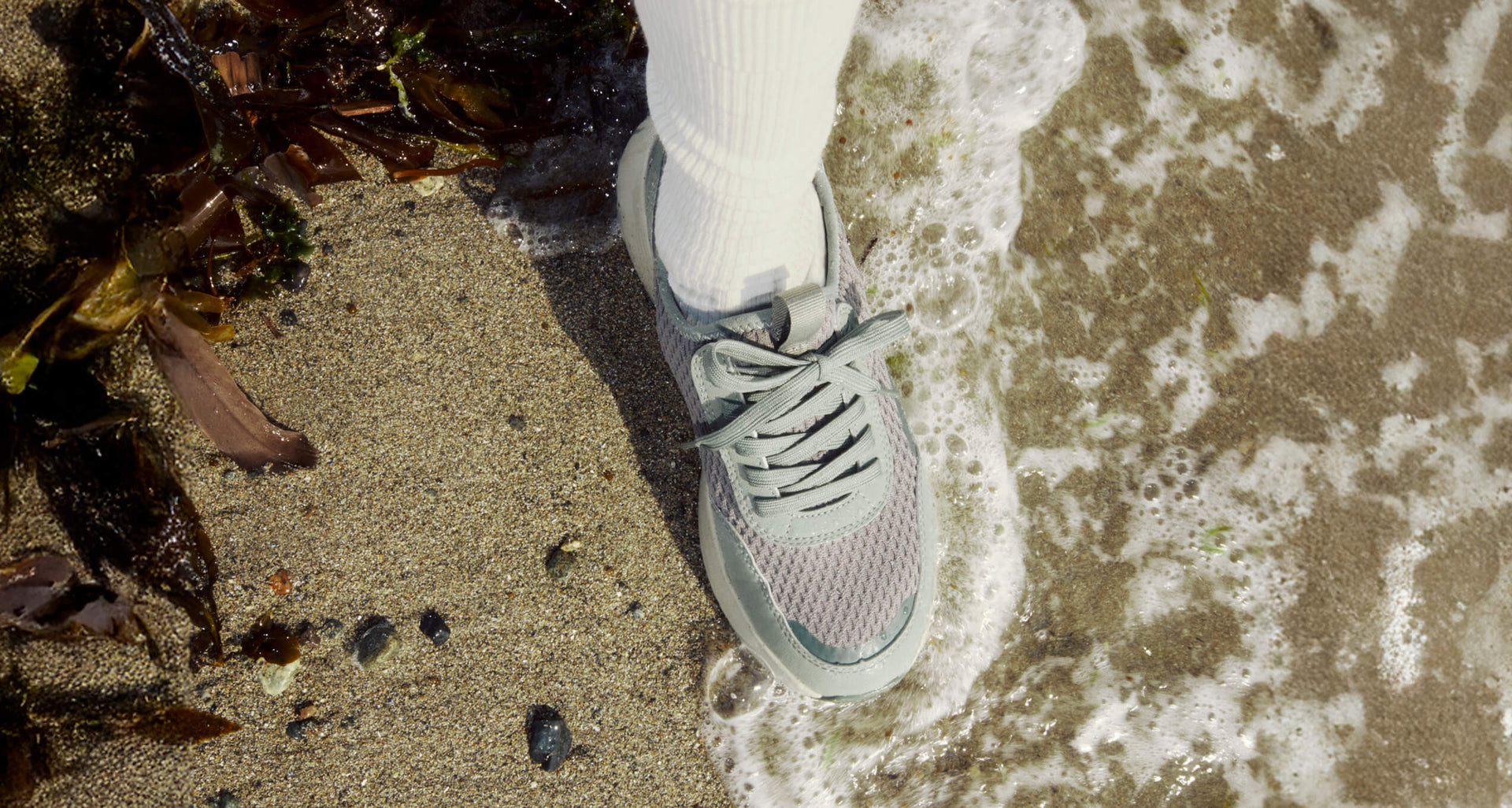 Foot wearing a gray sneaker stepping onto wet sand with seaweed.